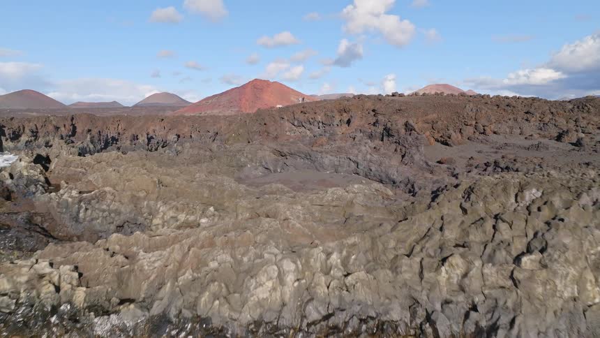 An aerial view rises over the dramatic volcanic coastline, revealing a rugged landscape of lava rock and distant volcanoes
