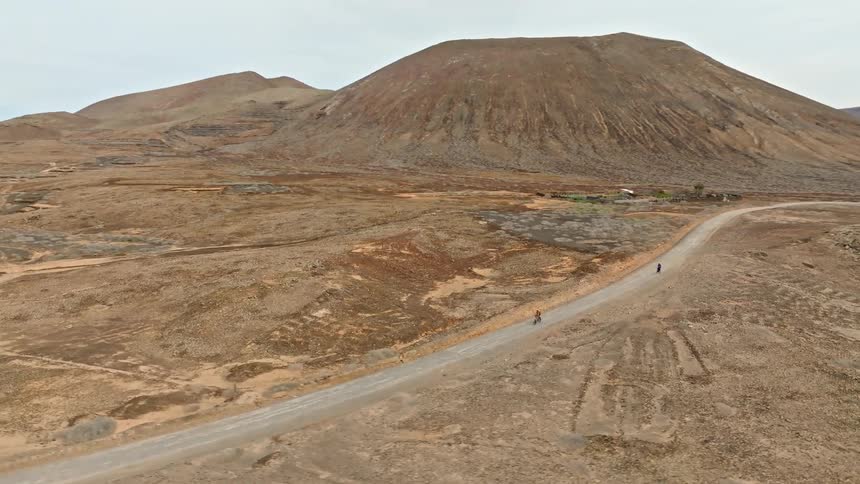 An expansive aerial view follows two cyclists as they ride through a vast, arid valley surrounded by volcanic mountains