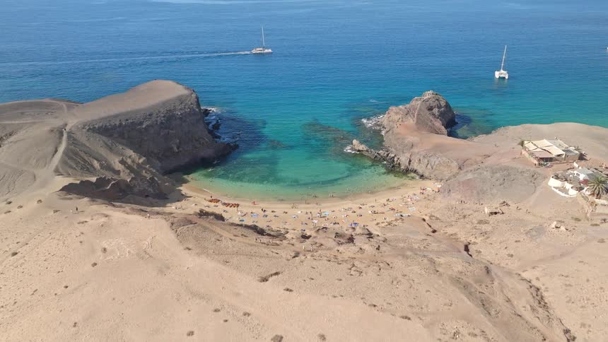 An aerial drone view flies towards a stunning secluded cove with turquoise water and a sandy beach full of people on a sunny day