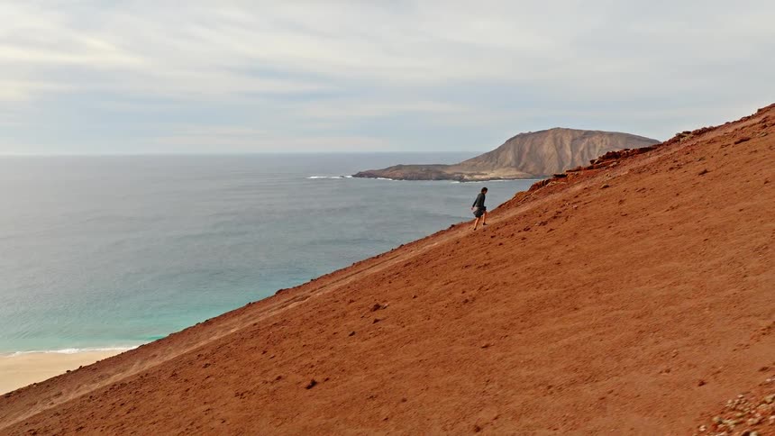 A lone adventurer hikes up a steep, red volcanic hill overlooking a beautiful, remote coastline
