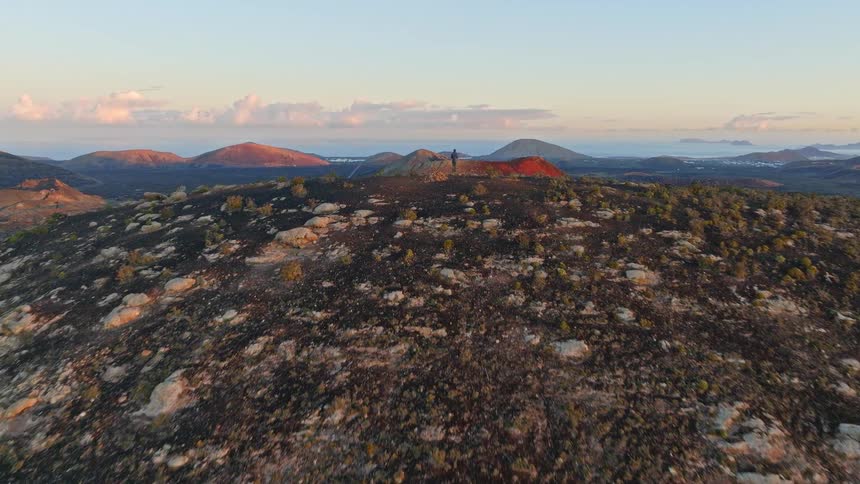 An aerial drone flies over a lone hiker on a volcanic summit, revealing a stunning panoramic view of the vast volcanic landscape below at sunrise