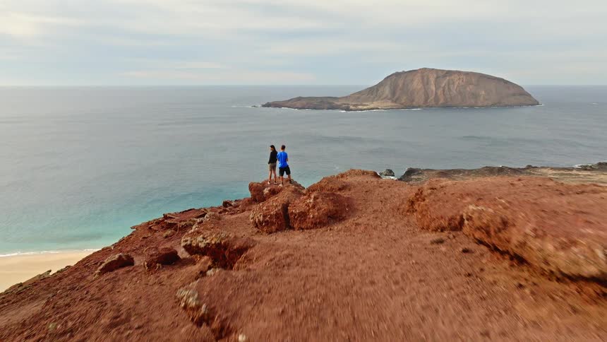 An adventurous couple stands on the edge of a dramatic red volcanic cliff, taking in the spectacular ocean and island views