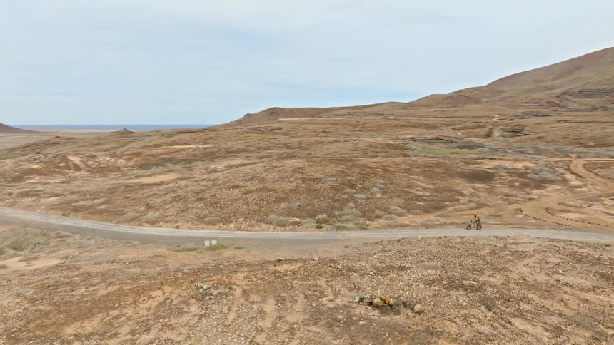 An aerial view follows a lone cyclist riding through a vast, barren volcanic landscape on a winding dirt road towards the distant ocean