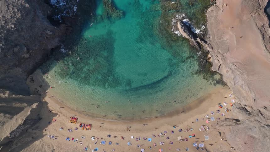Aerial top-down view zooms out from a stunning turquoise cove where tourists swim and sunbathe on a secluded sandy beach surrounded by rocky cliffs
