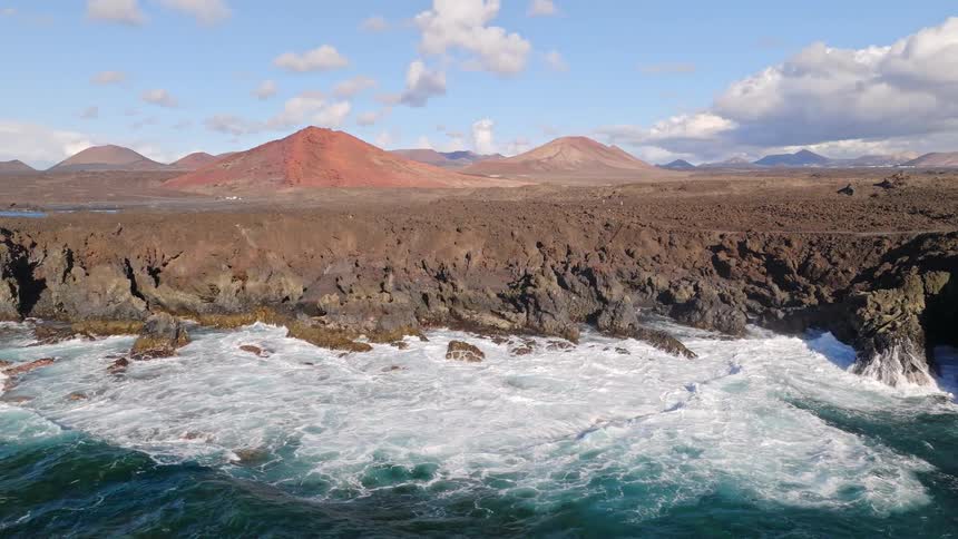 An epic aerial shot pulls back to reveal powerful turquoise waves crashing against a dramatic volcanic coastline with mountains in the background