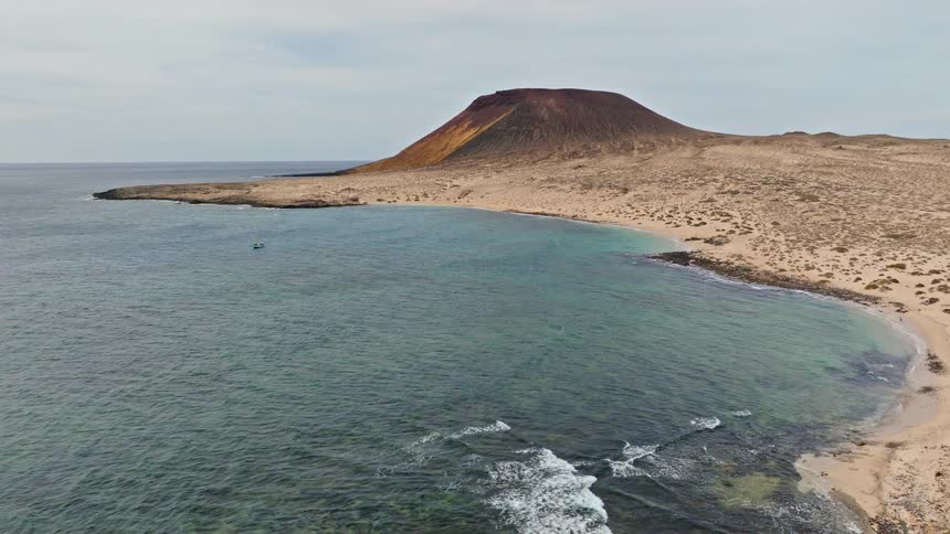 An ascending aerial view reveals a pristine turquoise bay and sandy beach next to a striking volcanic cone on a remote island