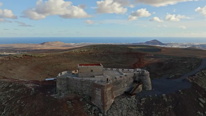 Aerial view of the historic Castillo de Santa Bárbara perched atop a volcanic crater, overlooking the vast landscape and ocean of Lanzarote