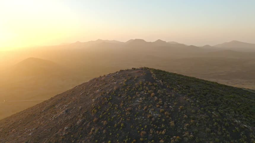 An epic aerial shot reveals hikers on a mountain summit, watching the golden sun rise over a hazy, volcanic landscape