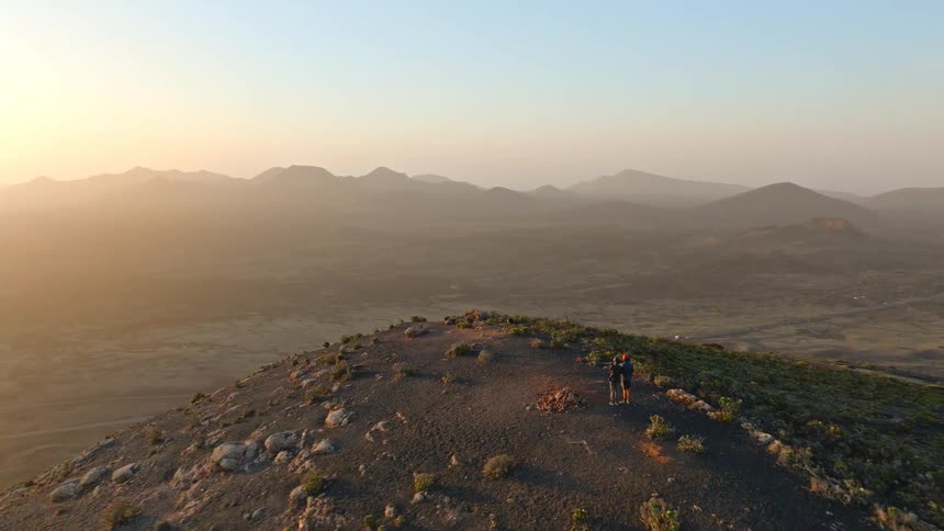 An aerial view shows a couple standing on a mountain peak, taking in the stunning sunrise over a vast volcanic landscape