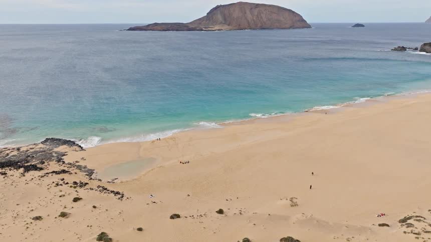 An expansive aerial view slowly pulls back from a turquoise shoreline to reveal a vast, secluded sandy beach and a distant rocky island