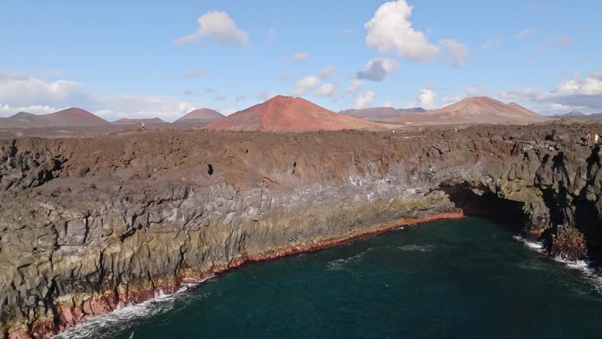 Aerial view rising over the dramatic volcanic coastline, revealing a stunning landscape of cliffs, sea caves, and distant volcanoes