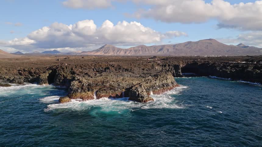An aerial drone shot pulls back to reveal the dramatic volcanic cliffs of Los Hervideros, Lanzarote, with powerful ocean waves crashing against the rocky shore