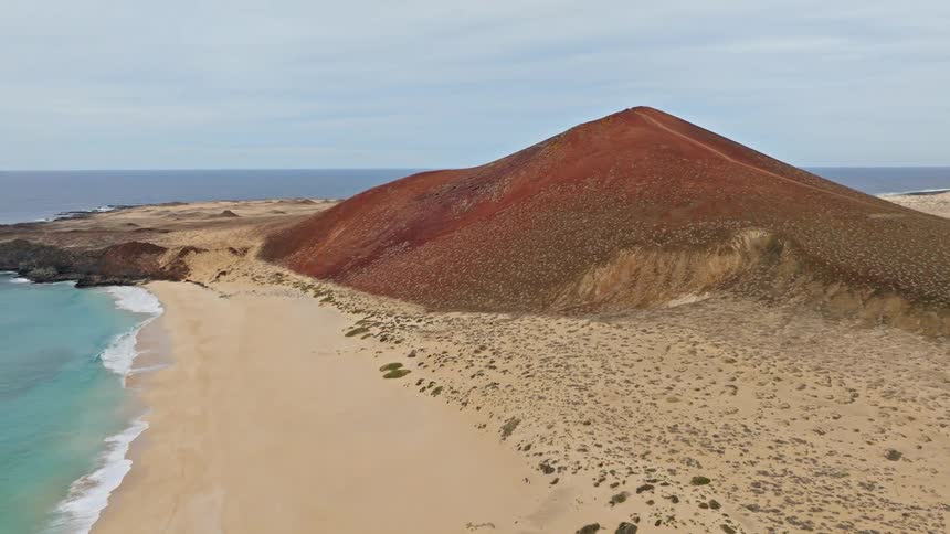 Aerial view pans over the dramatic red Montaña Bermeja volcano, revealing the beautiful golden sands of Playa de las Conchas on the island of La Graciosa