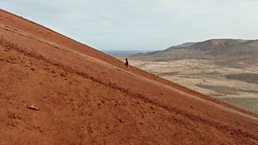 An adventurous hiker carefully descends the steep, red slope of a volcano with a vast, arid landscape and ocean in the background