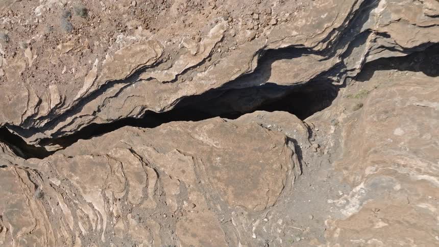 A dramatic top-down aerial shot slowly pulls away from a dark fissure in the earth, revealing the vast, layered rock formations of a dry canyon
