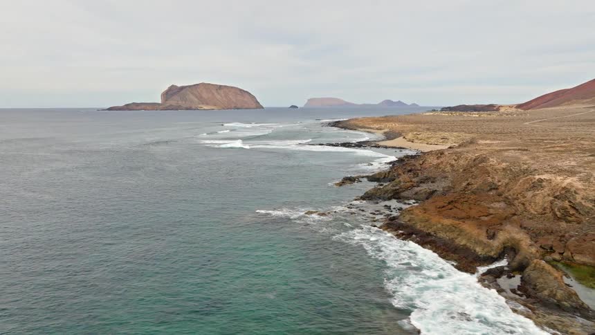 A cinematic aerial shot pulls back to reveal the dramatic, rocky volcanic coastline of Lanzarote, with the red Montaña Bermeja and the Chinijo Archipelago in the background