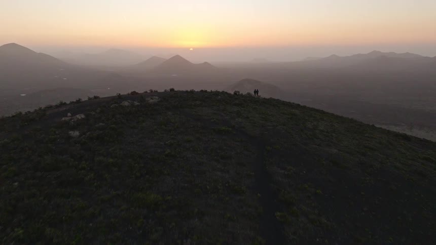 A stunning aerial shot rises over a silhouetted couple on a mountaintop, revealing a vast volcanic landscape during a golden sunrise