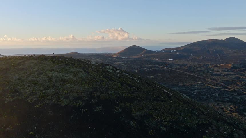 An epic aerial shot reveals two hikers on a volcanic summit at sunrise, overlooking a vast landscape of craters and lava fields