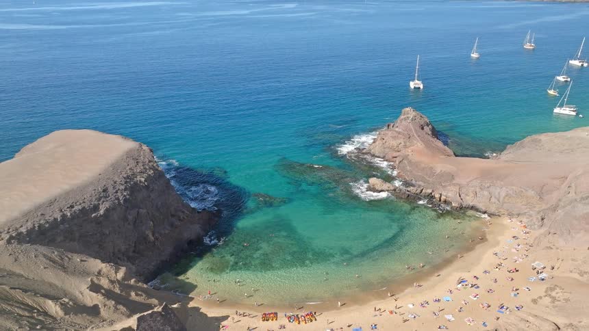 Aerial view pulling back from a stunning secluded cove with a golden sand beach, clear turquoise water, and sailboats on the horizon