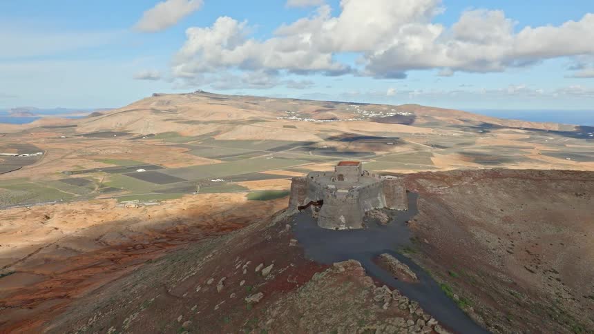 An aerial approach to the historic Santa Barbara Castle perched on a volcanic crater in Teguise, Lanzarote, revealing the expansive island landscape