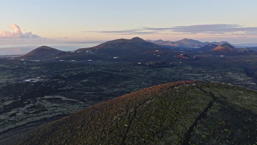 A rising aerial shot reveals a stunning volcanic landscape with multiple craters at sunrise, with the ocean visible on the horizon