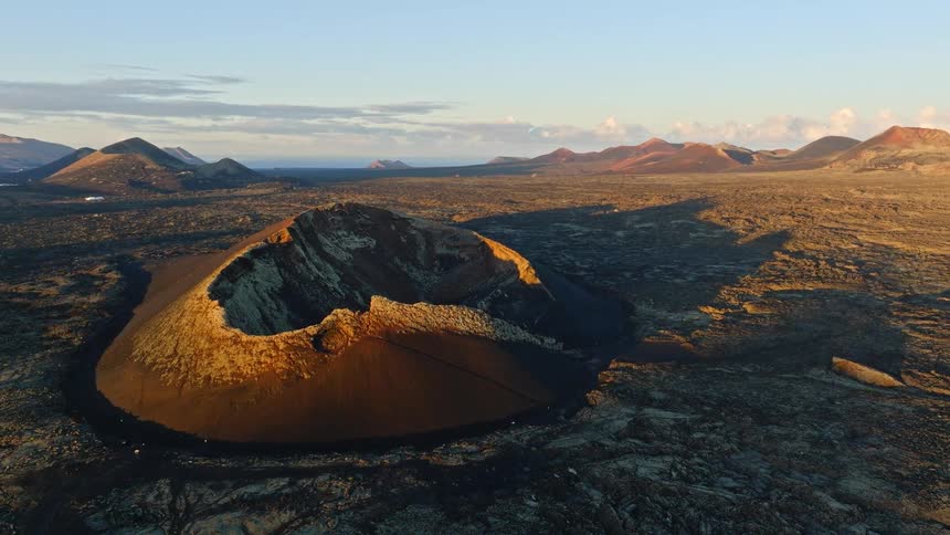 An epic aerial drone shot circles the majestic El Cuervo volcano crater in Lanzarote, illuminated by the warm light of a beautiful sunset