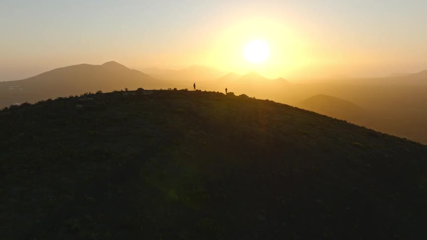 An aerial view pulls back to reveal two hikers silhouetted on a mountain peak enjoying the golden sunrise over a vast landscape
