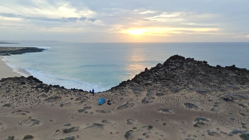 A dramatic aerial shot orbits a lone camper with a tent on a volcanic ridge, revealing a stunning deserted beach at sunset