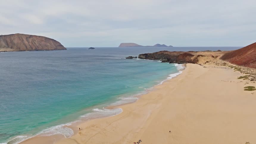 A stunning panoramic aerial view reveals a vast, secluded golden sand beach with turquoise waters, rolling waves, and distant volcanic islands