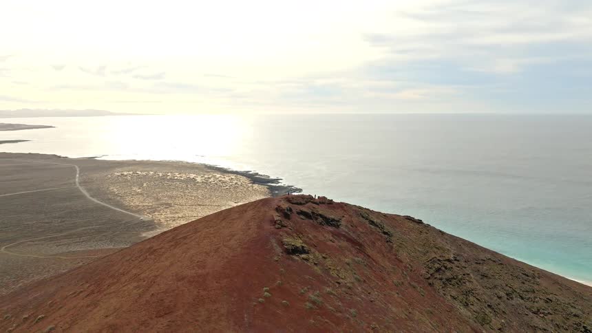 Aerial pull-back shot reveals a solitary person standing atop a red volcanic cone, overlooking a stunning island coastline and the vast ocean