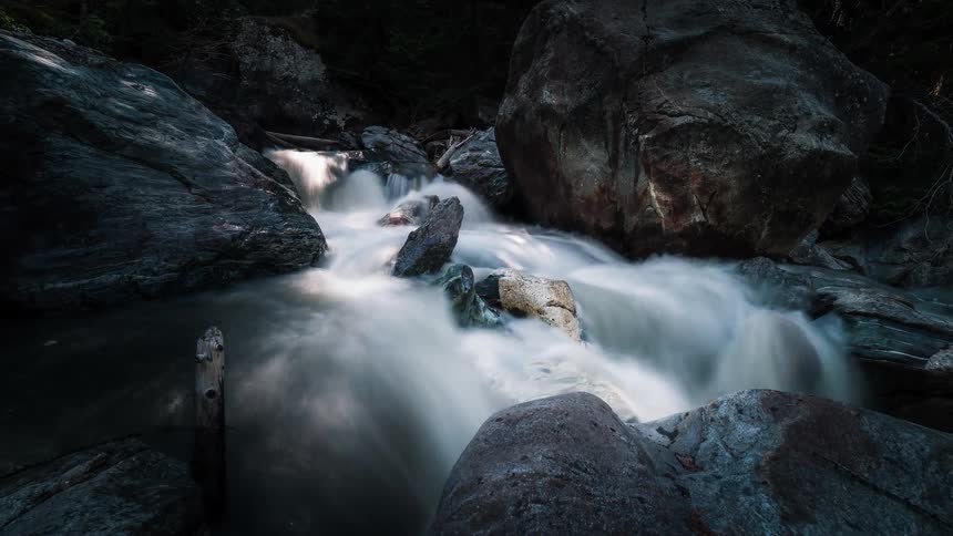 A mystical long exposure time-lapse captures a river flowing over large boulders in a dark forest as sunlight moves across the water