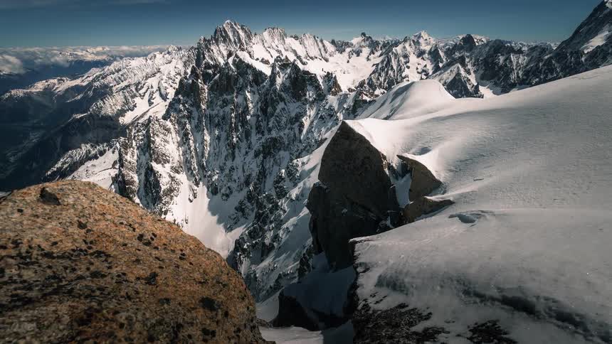 A slow pullback shot reveals a vast and majestic snow-covered mountain range on a bright, clear day