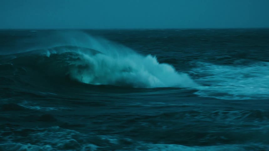 A powerful ocean wave crests and crashes in dramatic slow motion under the dark, moody light of dusk