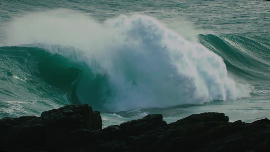 An immense turquoise ocean wave crashes in dramatic slow motion with dark rocks silhouetted in the foreground