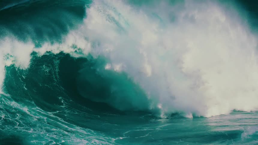 An immense teal-colored ocean wave dramatically curls and crashes in breathtaking slow motion