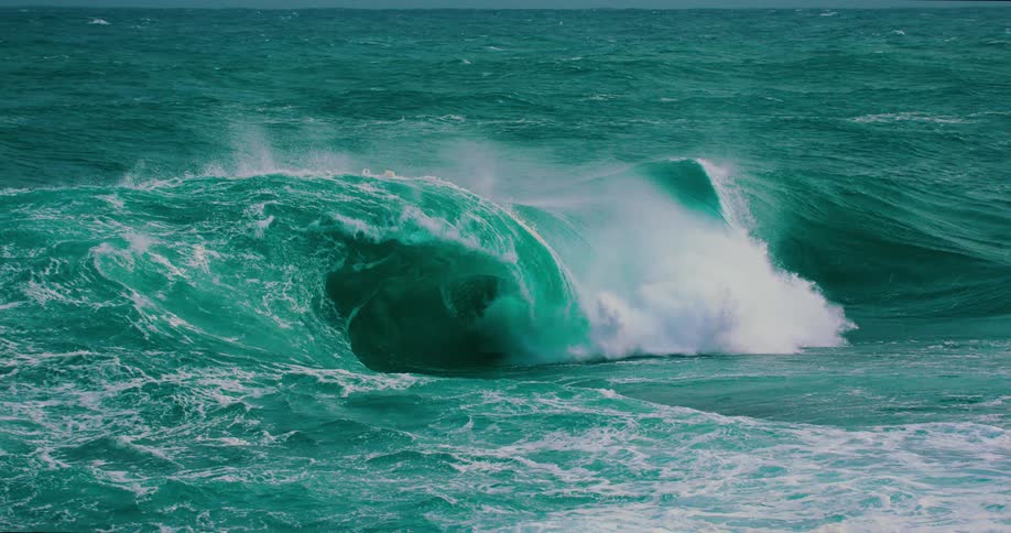 A powerful turquoise ocean wave curls and crashes in dramatic slow motion, creating a plume of white sea foam