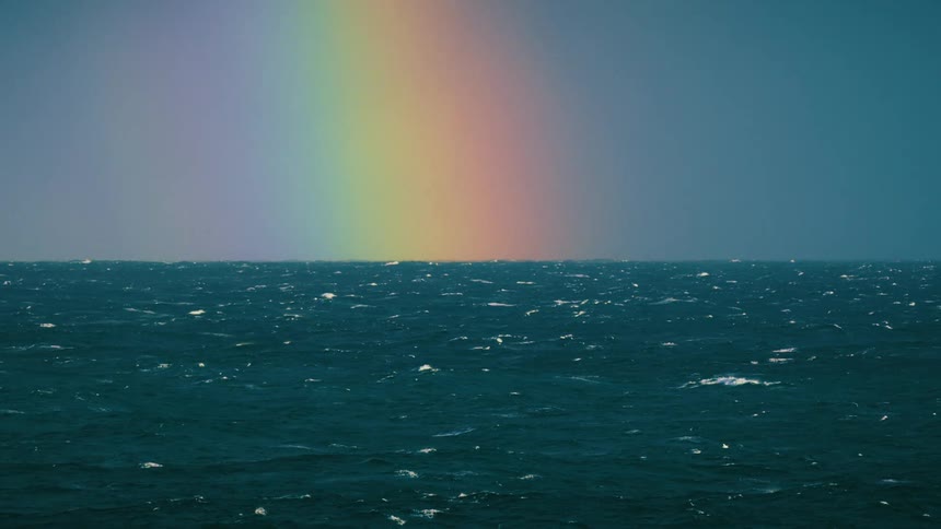 A vibrant rainbow emerges from the horizon, arching over a deep blue, choppy ocean under a dramatic sky