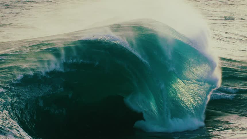 A giant, powerful ocean wave builds and crashes in dramatic slow motion, its green-blue water illuminated as it curls into a massive barrel