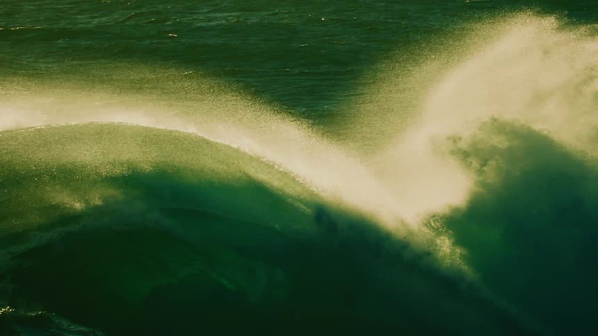 A powerful ocean wave curls in dramatic slow motion, its green surface illuminated by backlight as mist blows from its crest