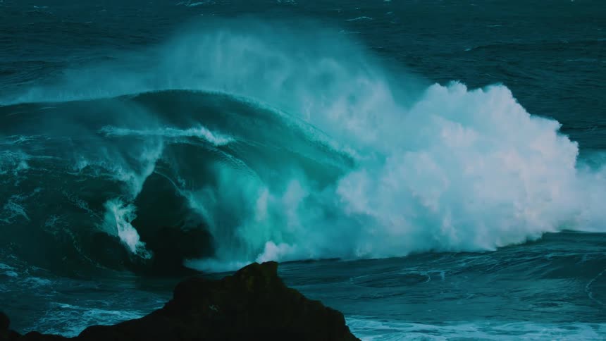 A massive, powerful ocean wave with teal-colored water curls and crashes in dramatic slow motion