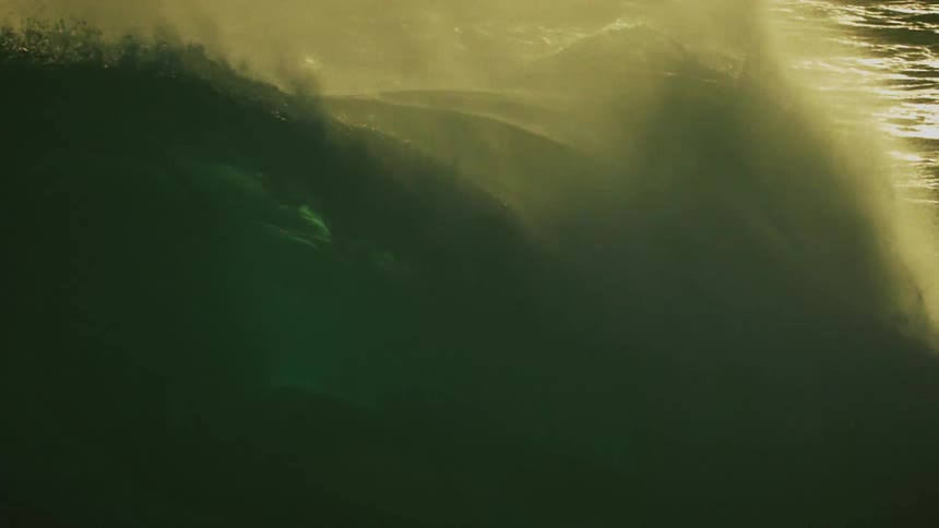 A massive, dark wave crests and breaks in dramatic, backlit slow motion