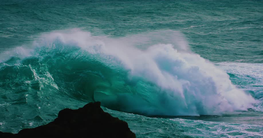 A dramatic slow-motion shot captures a massive turquoise ocean wave as it curls and crashes with immense power