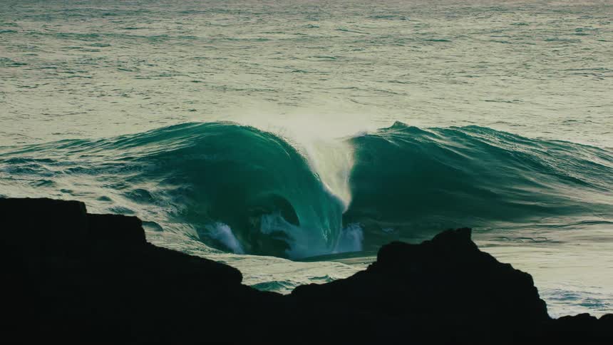 An epic, turquoise ocean wave crests and crashes in super slow motion as seen from a rocky coastline