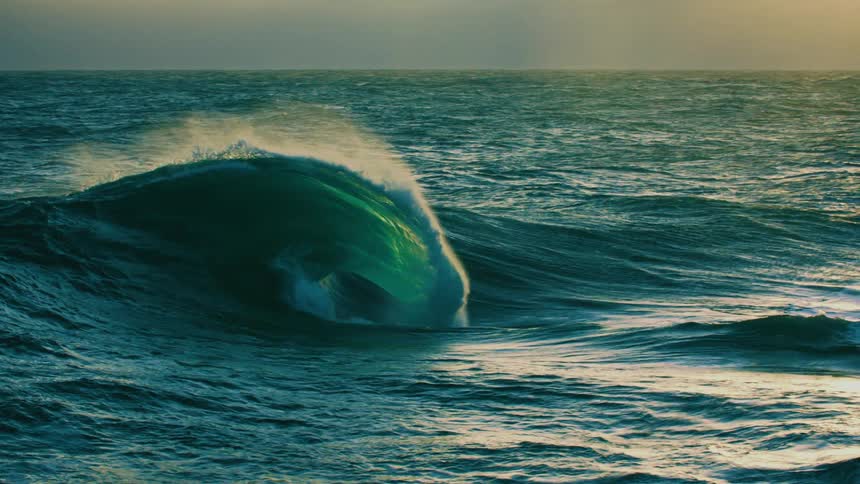 A powerful turquoise ocean wave forms and breaks in dramatic slow motion as the sunlight illuminates it from behind