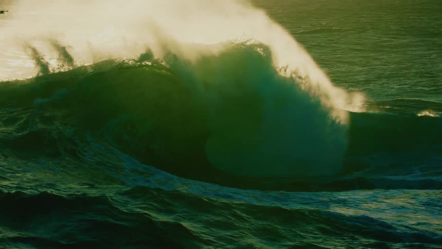 A massive, powerful ocean wave builds and crashes in dramatic slow motion, backlit by the golden sun