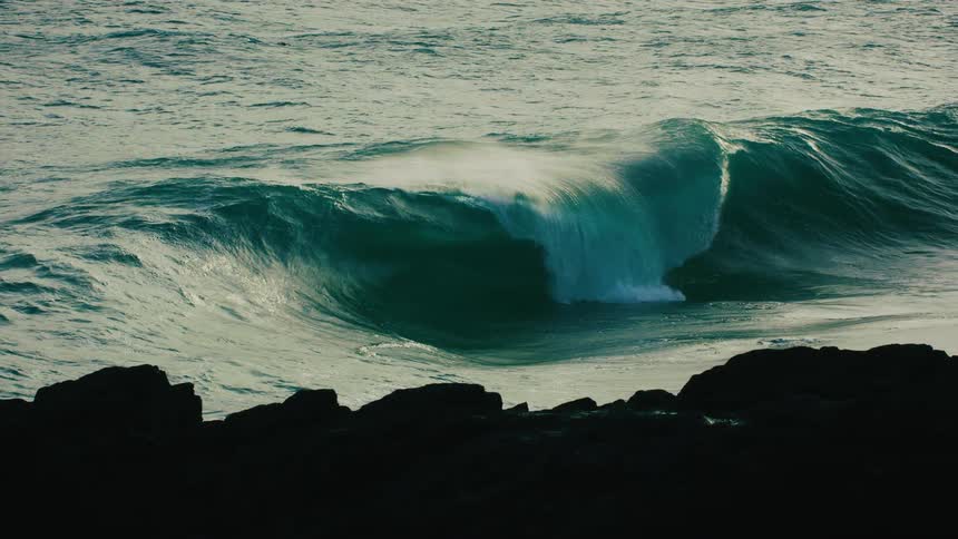 A powerful turquoise ocean wave builds and crashes in epic slow motion against a dark, rocky shoreline
