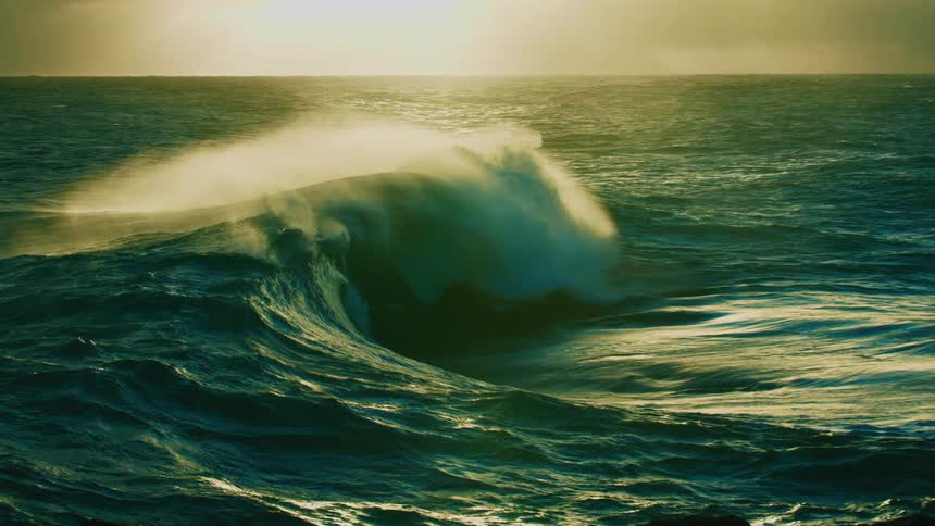 A massive ocean wave crests and breaks in dramatic slow motion with the golden sunlight of a sunset or sunrise in the background