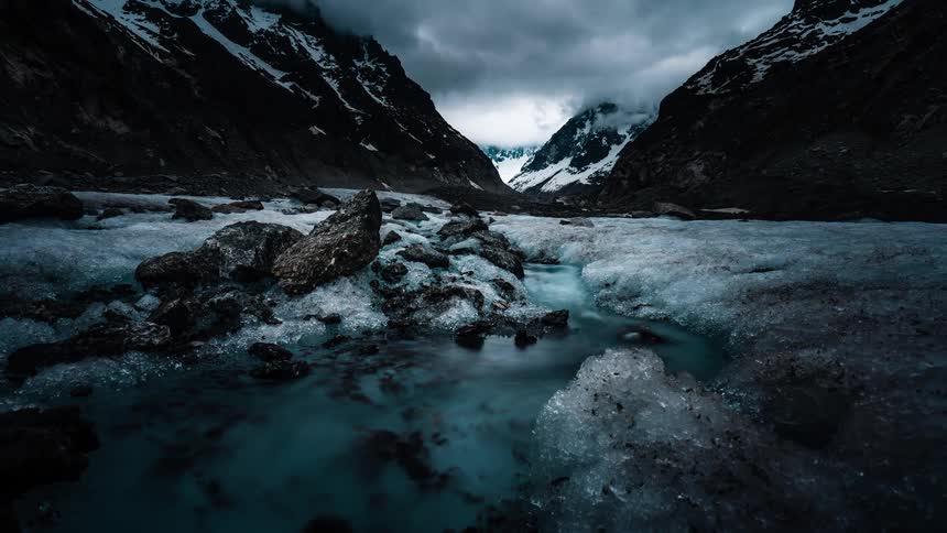 A glacial river of turquoise water flows through an icy valley between dark, snow-capped mountains under a stormy sky