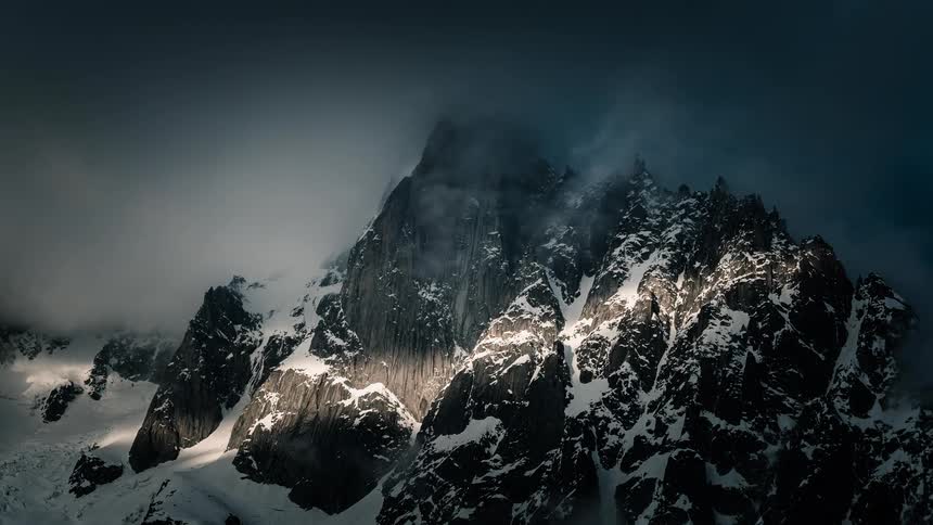 Sunlight pierces through dramatic, swirling clouds to illuminate a majestic snow-covered mountain peak