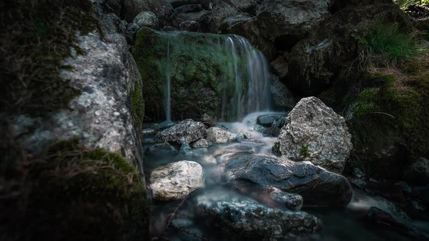 A serene long-exposure timelapse reveals a small, enchanting waterfall cascading over mossy rocks in a tranquil forest stream